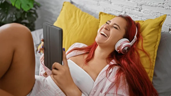 Cheerful young redhead woman cosily lying in bed, indulging in digital entertainment, blissfully watching a video and smiling inside her beautiful bedroom
