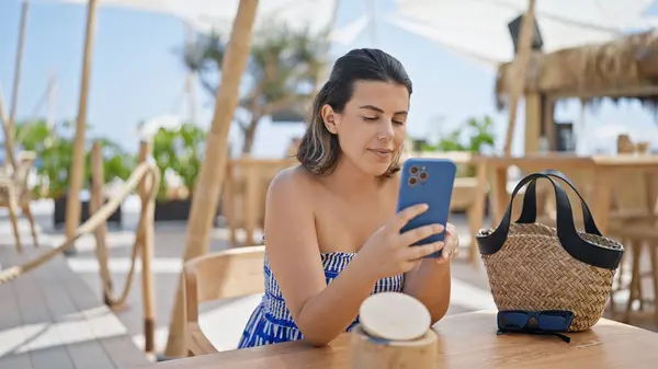 Young hispanic woman using smartphone sitting on the table at sunny restaurant terrace