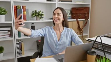 Işıl ışıl genç İspanyol kadın ofis işi sırasında akıllı telefonuyla selfie çekiyor. Şık iş yerindeki başarılı iş hayatına dair bir fotoğraf.