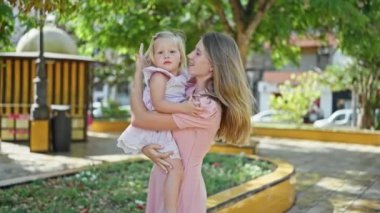 Kendine güveni tam bir anne, kollarını kızına dolamış, neşeyle gülüyor, güneşli parkta duruyor, pozitif enerji ve mutluluk saçıyor.