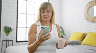 Yatakta rahatlarken gülümseyen orta yaşlı, kendine güvenen sarışın bir kadın. Kahve içiyor ve yatak odasının rahatlığında akıllı telefonuyla mesajlaşıyor.