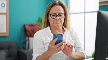 Orta yaşlı İspanyol iş kadını. Ofiste bilgisayar kullanıyor. Akıllı telefondan konuşuyor.