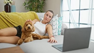 Dizüstü bilgisayarında film izleyen beyaz bir kadın. Evdeki kanepede yatıp gülümsüyor.