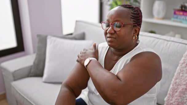 严重的非洲裔美国女人 戴着眼镜和辫子 坐在舒适的客厅沙发上 在家里肩膀受伤 很不舒服 — 图库视频影像