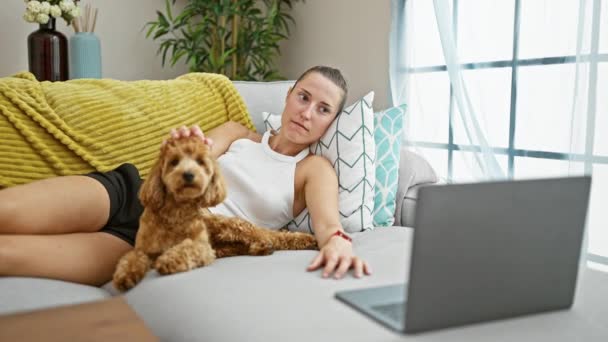自宅で笑顔のソファーに横たわるラップトップで映画を見ている犬を持つ若い白人女性 — ストック動画