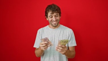 Arjantin Pesos banknotlarıyla servetini güvenle sayan genç adam, canlı kırmızı bir arka planda izole edilmiş.