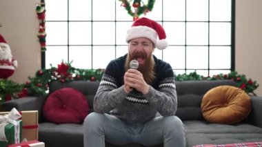 Genç kızıl saçlı adam evdeki Noel süslemesinin yanında kanepede şarkı söylüyor.