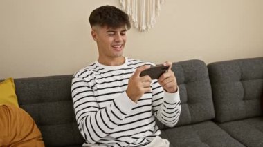 Mutlu İspanyol genç erkek oyuncu video oyunu oynarken zaferi kutluyor, gülümseyen adam oturma odasındaki kanepe konforunda akıllı telefondan kazanmayı seviyor, evde eğlenceli teknoloji kutlaması.