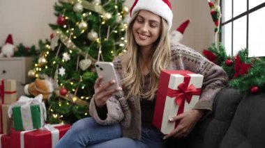 Genç sarışın kadın elinde akıllı telefon hediyesiyle evde Noel ağacının yanında oturuyor.