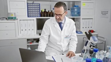 Endişeli, orta yaşlı, gri saçlı bilim adamı laboratuvarda dizüstü bilgisayarla çalışırken üzgün.