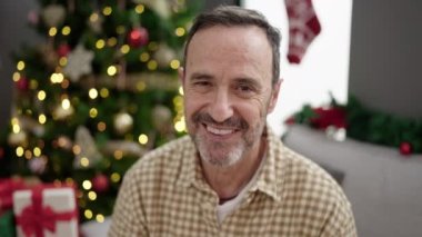 Orta yaşlı bir adam evde Noel ağacının yanında kendinden emin bir şekilde oturuyor.
