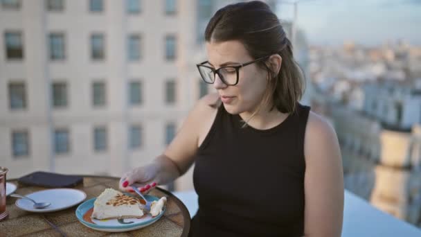 おいしい脱出 メガネの美しいヒスパニック女性 おいしいチーズケーキのスライス 街の空を見下ろす現代的な屋上テラスに座って — ストック動画
