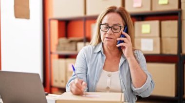 Orta yaşlı İspanyol kadın ecommerce iş işçisi pakete yazı yazıp ofisteki akıllı telefondan konuşuyor.