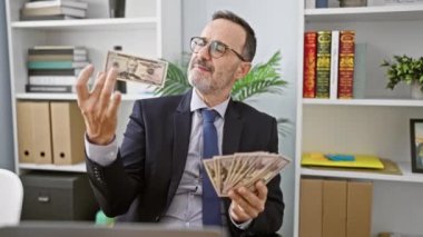 Gri saçlı neşeli orta yaşlı bir adam, gülümseyen bir iş adamı, işyerindeki ofisine neşeyle dolar fırlatıyor.