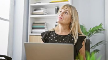 Orta yaşlı, sarışın bir kadın ofis bilgisayarında işini yaparken derin düşüncelere dalmış zarafet ve profesyonel başarı havası yayıyor.