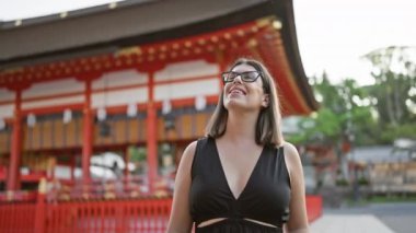 Kendinden emin bir şekilde Fushimi Inari Taisha 'ya poz veriyor, gözlüklü güzel bir İspanyol kadın, geniş bir şekilde gülümsüyor, etrafına bakıyor, büyüleyici Kyoto tapınağı çevresinden büyüleniyor..