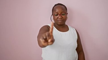 Dayanıklı Afro-Amerikan kadın, artı boyut, gündelik tarzda duruyor, ciddi bir ifadeyle 'hayır' diyor ve izole edilmiş pembe arka planda parmak yükseliyor..