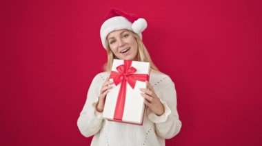 Sarışın, genç bir kadın, Noel şapkası takıyor ve kırmızı arka planda hediyeler tutuyor.