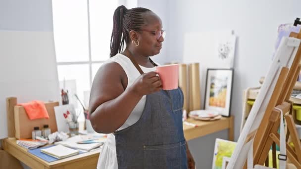 アフリカ系アメリカ人女性アーティスト コーヒーのカップで思慮深く立って 彼女のアートスタジオで彼女の絵を見て — ストック動画