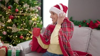 Genç sarışın kadın müzik dinliyor. Evde Noel ağacının yanında kahve içiyor.