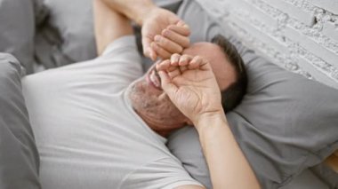 Gri saçlı, rahat bir sabah geçiren orta yaşlı bir adam, rahatlatıcı bir yatak odasında yarı uyanık bir şekilde uzanmış, kapalı dairesindeki yastıkla kaplı yatağına gözlerini sürterek