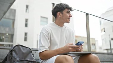 Günışığı genç İspanyol üniversite öğrencisi, şehir kampüsünün merdivenlerinde otururken telefonuyla neşeli bir şekilde mesajlaşıyor, manyetik gülümsemesiyle kendine güvenini yayıyor.