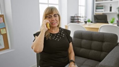 Zarif sarışın, orta yaşlı kadın patron, akıllı telefonuyla ciddi bir iş konuşmasına dalmış, iş yerinde kapalı bir yerde çalışıyor..