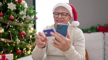 Orta yaşlı, kır saçlı, akıllı telefon ve kredi kartıyla alışveriş yapan bir adam evdeki Noel ağacının yanında koltukta oturuyor.
