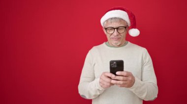 Orta yaşlı, gri saçlı, Noel şapkası takan, izole edilmiş kırmızı arka planda akıllı telefon kullanan bir adam.