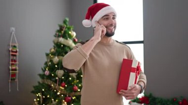 Genç Arap adam elinde hediyeyle akıllı telefondan konuşuyor. Yılbaşı ağacının yanında duruyor.