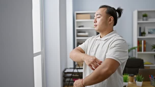 ビジネスワーカーの若い中国人男性は オフィスの窓を一目見て投げます 立って 腕を折って 彼は職場の問題を考えて 屋内で問題を抱えているように見えます — ストック動画
