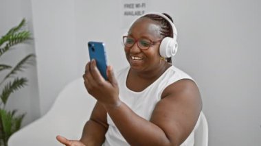 Afro-Amerikan kadın, neşeyle gülümsüyor, bekleme odasında bir iş videosu var, rahatça oturuyor, odaya ruhsal ritim ekliyor.