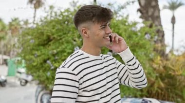 Kendine güvenen genç İspanyol adam, güneşli parkın yeşilinin altında dimdik duruyor, gerçekten gülümsüyor. Havalı görünmesi, akıllı telefonuyla konuşması, mutluluk saçması....