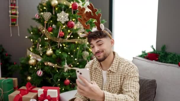 若いアラブの男は自宅でクリスマスツリーに座ってスマートフォンでセルフィーを作ります — ストック動画