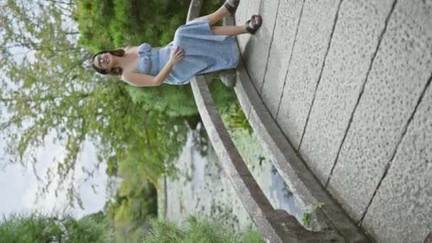 日本の公園の橋の上に座って笑顔の眼鏡をかけた美しい若いヒスパニックの女性 — ストック動画