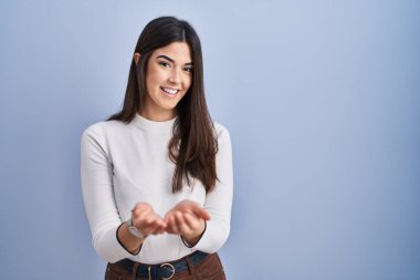 Mavi arka planda duran esmer kadın avuçlarını birbirine uzatarak gülümsüyor ya da el hareketi yapıyor. Bekleme ve koruma 