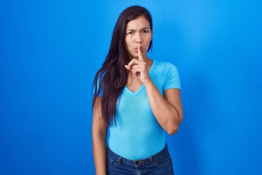 Mavi arka planda dikilen genç İspanyol kadın, parmakları dudaklarında sessiz olmak istiyor. sessizlik ve gizli konsept. 