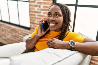 Afro-Amerikalı kadın akıllı telefonda konuşuyor. Evdeki kanepede oturuyor.