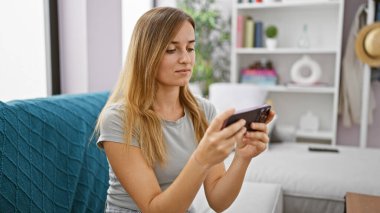 Genç sarışın kadın evdeki kanepede oturup video oyunu oynamaya odaklanmış.