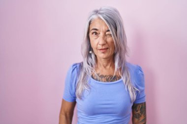 Pembe arka planda dövmeleri olan orta yaşlı bir kadın kameraya seksi bir ifadeyle bakıyor, neşeli ve mutlu bir yüzle.. 