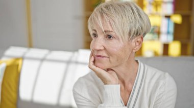Orta yaşlı sarışın bir kadın, rahat bir ifadeyle koltukta oturuyor ve evine bakıyor.