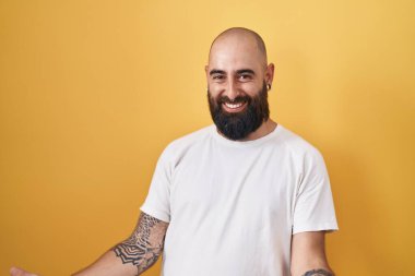Sarı arka planda dikilen sakallı ve dövmeli genç İspanyol adam. Kolları açık gülümsüyor. Dostça karşılama, olumlu ve kendinden emin selamlar. 