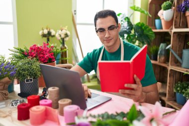 Young hispanic man florist using laptop reading book at flower shop