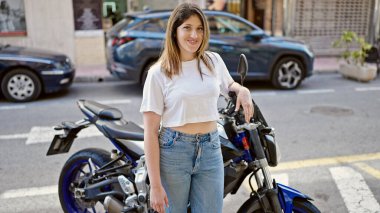 Genç sarışın kadın gülümsüyor. Motosikletin yanında duruyor. Kollarını kavuşturup sokakta el hareketi yapıyor.