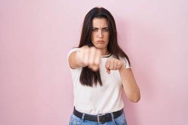 Genç esmer kadın pembe arka planda duruyor. Yumruk yumruğa dövüşmek, agresif ve öfkeli saldırı, tehdit ve şiddet. 