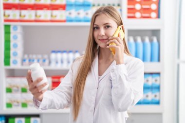 Kafkas kadın eczacı elinde ilaç şişesiyle eczanede akıllı telefondan konuşuyor.