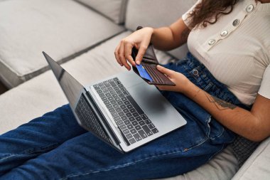 Evdeki kanepede oturan, dizüstü bilgisayar kullanan genç ve güzel bir İspanyol kadın.