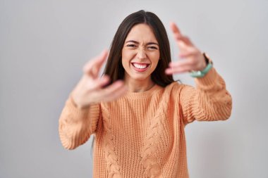 Beyaz arka planda duran esmer kadın öfkeyle bağırıyor, elleri boğazlamaya çalışıyor, deli gibi bağırıyor. 