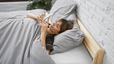 Yatakta uzanmış akıllı telefon kullanan genç, güzel İspanyol kadın.