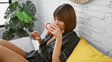 Rahat, genç ve güzel İspanyol kadın, kısa saç stiliyle rahatlamış, pijamalarıyla yatakta rahat bir şekilde oturuyor, sıcak yatak odasında sabah kahvesinin tadını çıkarıyor ve mesajlaşıyor.